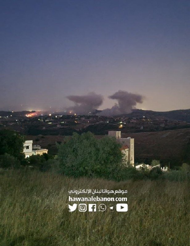 A twilight landscape shows distant smoke plumes rising above a mountainous horizon with scattered town lights, while a stone building and trees stand in the foreground beneath a logo reading "hawanalebanon.com."