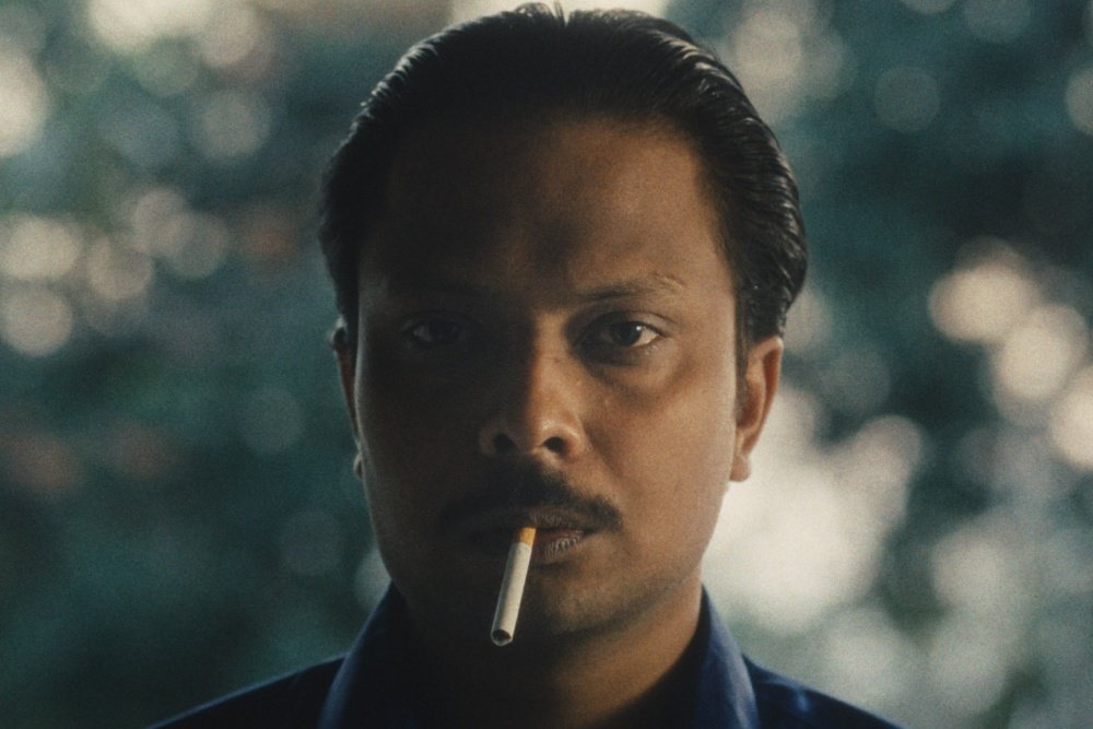 A close-up image shows a man with slicked-back hair and a mustache holding an unlit cigarette in his mouth, set against a blurred green background.