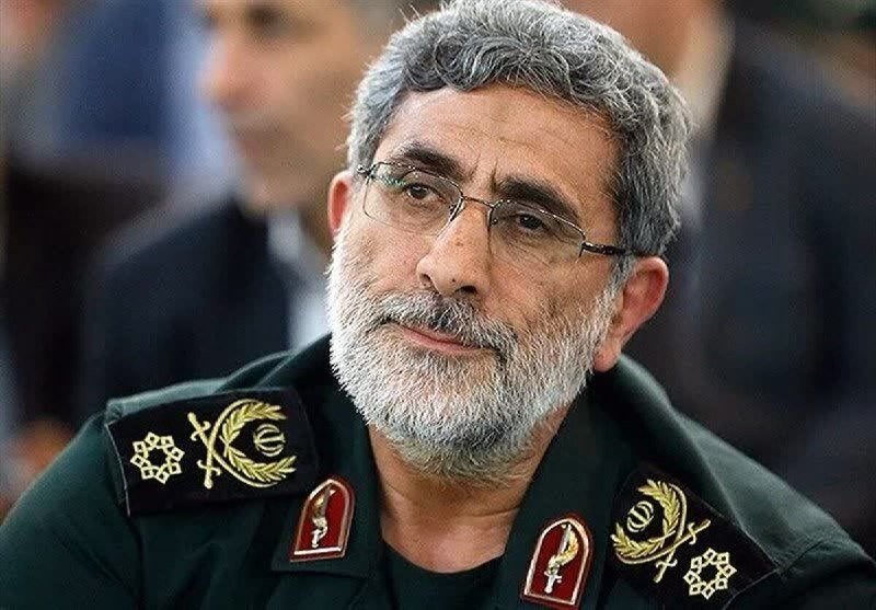 A gray-haired, bearded man in a green military uniform with insignia and shoulder patches gazes forward, with a blurred figure visible behind him.