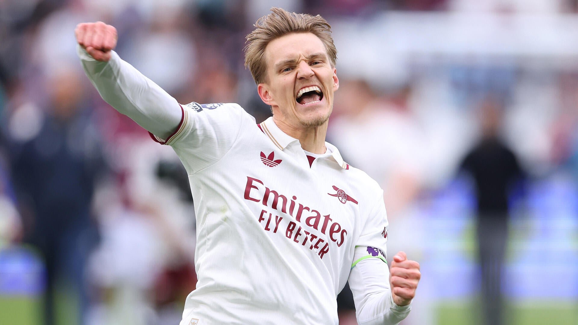 A blond male soccer player in a white Arsenal jersey shouts with a raised fist, celebrating on a pitch before a blurred crowd.