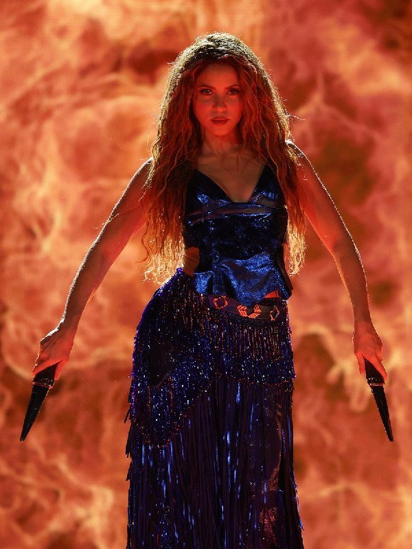 A woman with long red hair, wearing a blue sequined top and fringed skirt, holds pointed objects in both hands against a fiery orange background.