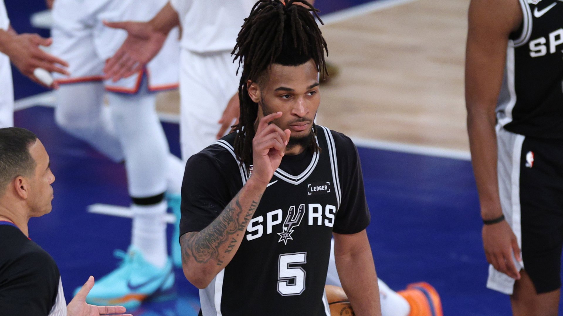 A basketball player wearing a black San Antonio Spurs jersey with the number 5 stands on a court, raising one finger while holding a basketball between his legs.