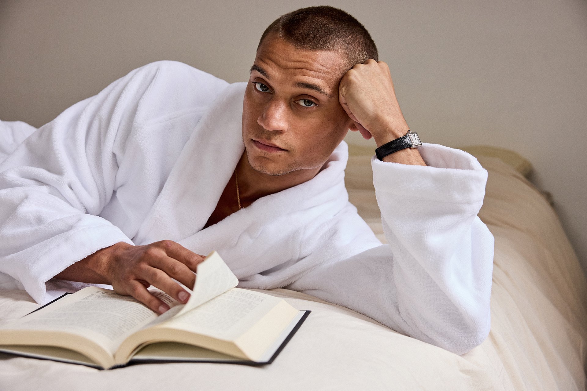A man in a white bathrobe reclines on a bed, holding an open book while wearing a wristwatch.