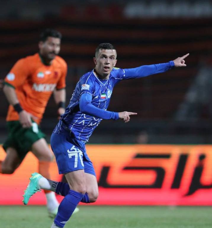 A soccer player in a blue uniform celebrates by pointing his finger while running, with an opposing player in an orange jersey visible behind him on the field.