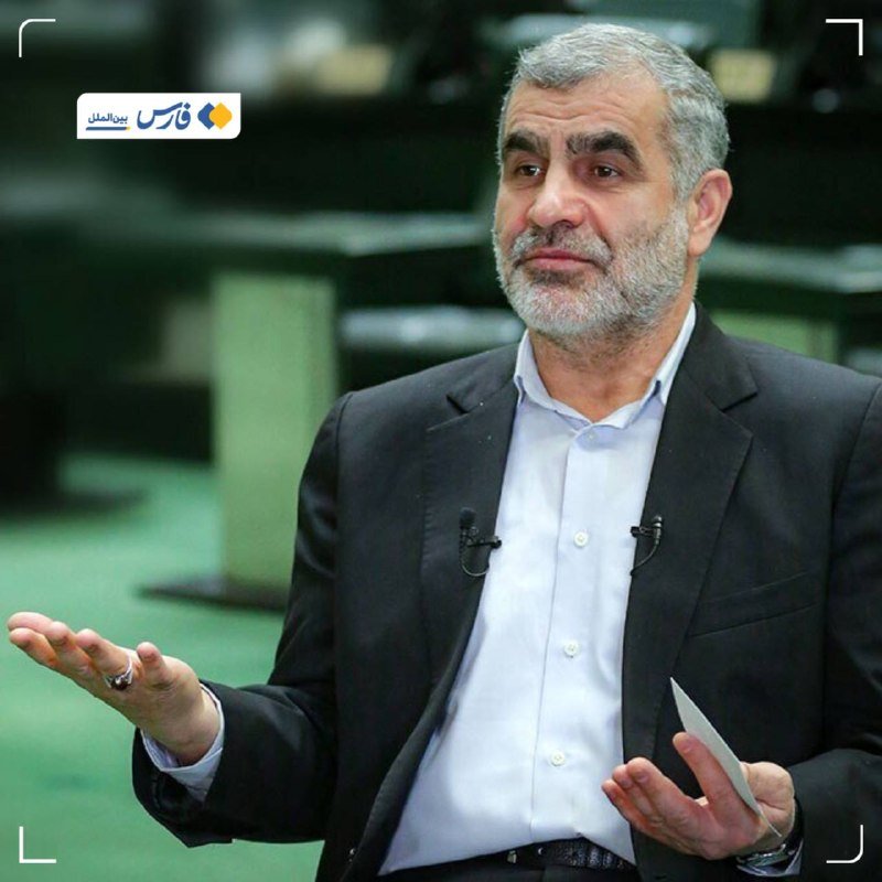 A gray-bearded man in a black suit and light blue shirt gestures with his hands while speaking, holding papers, in a green-toned parliamentary chamber setting with a news channel logo.