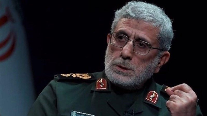 A gray-haired, bearded man in a green military uniform with insignia and glasses gestures with his hand during what appears to be an interview, with a partially visible flag behind him.