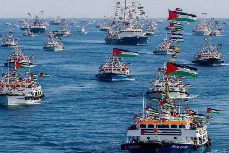 A large flotilla of boats sails on open water, many flying Palestinian flags, with one prominent vessel in the foreground displaying an orange "Wave to Gaza" banner.