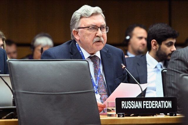 A gray-haired man in a suit and glasses speaks at a desk with a "RUSSIAN FEDERATION" placard in a formal assembly setting.
