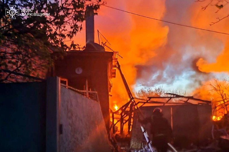 Large flames engulf a multi-story building at night, with two figures on the roof and a firefighter climbing a ladder amid smoke and debris.