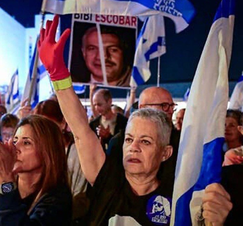 A crowd of demonstrators holds up a poster bearing a man's photo with Hebrew text and waves blue-and-white flags during a nighttime rally.