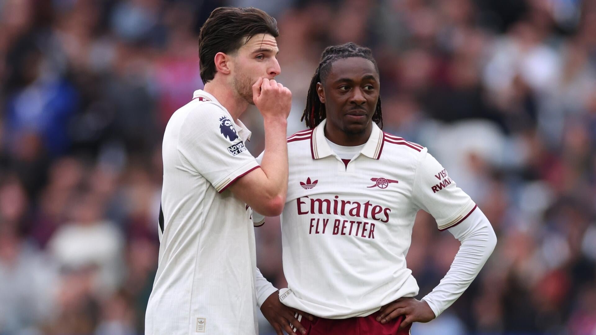 Two soccer players in white Arsenal jerseys stand on the field during a match, one with his hand near his mouth and the other with hands on his hips, against a blurred crowd.