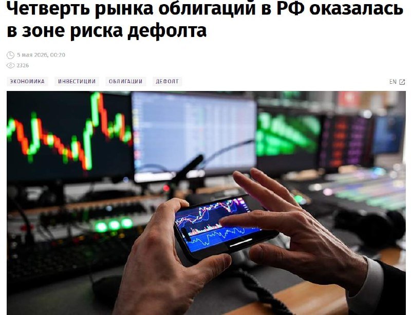 A person holds a smartphone displaying a stock chart, tapping the screen with one hand, while multiple monitors showing financial charts are visible in the background.