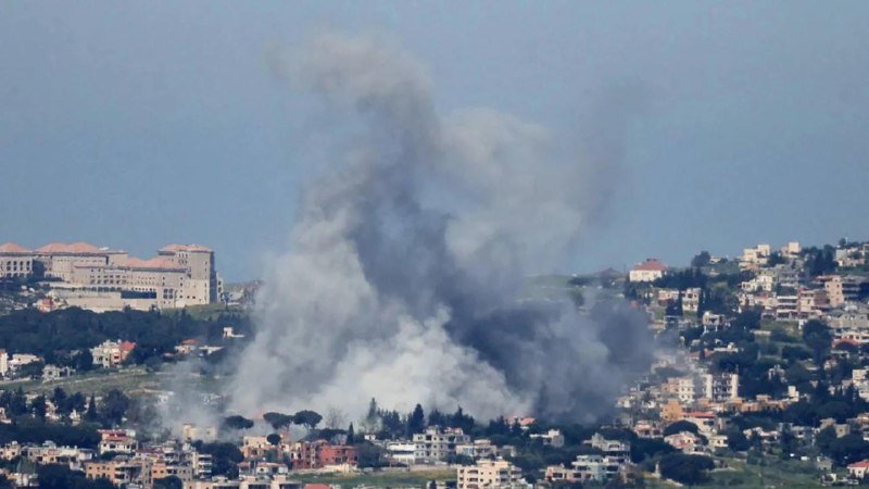 Large plumes of gray and white smoke rise above a hillside town with densely packed buildings under a hazy sky.