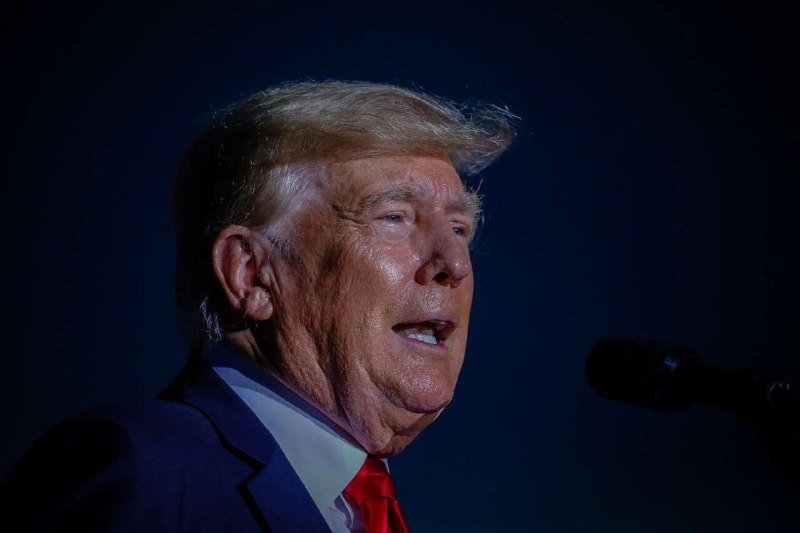 A man with blond hair in a dark suit and red tie speaks into a microphone against a dark blue background.