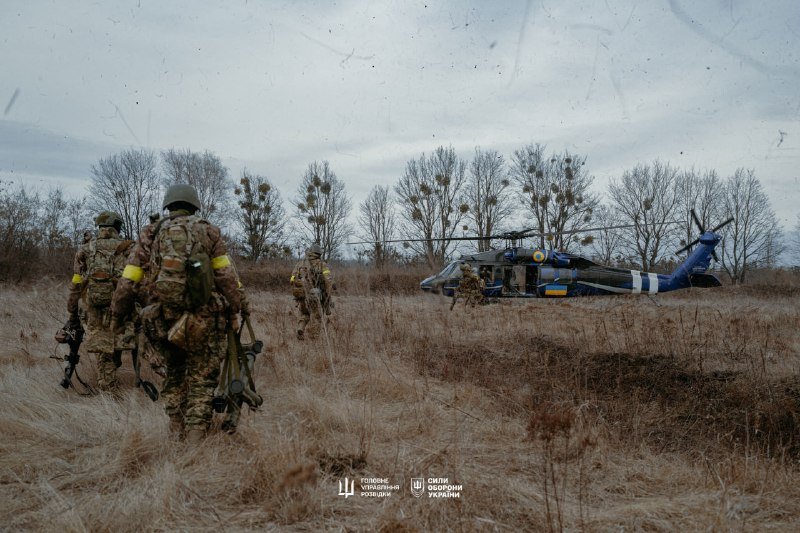 Soldiers in camouflage gear with yellow armbands walk through a grassy field toward a blue and yellow helicopter near bare trees under an overcast sky.