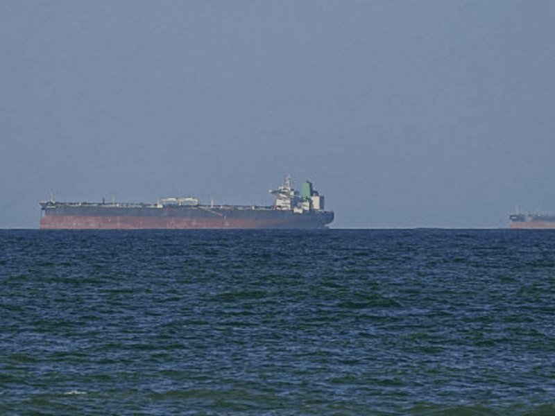 A large oil tanker floats on choppy blue-green ocean waters under a hazy gray sky, with another ship visible on the distant right horizon.