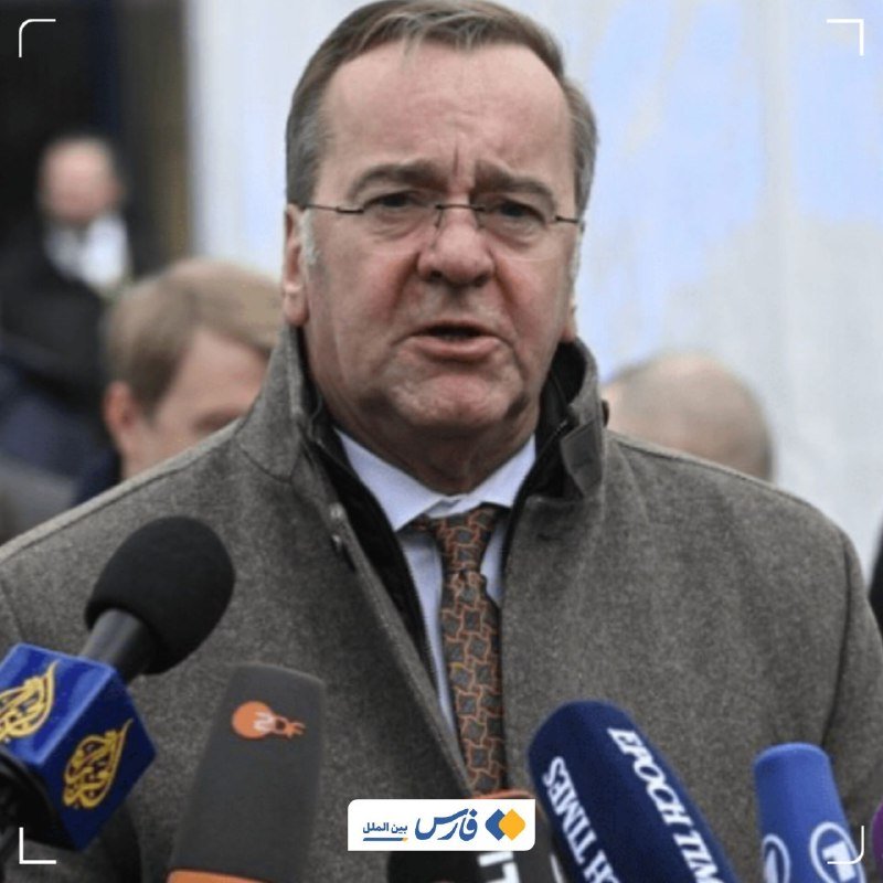 A bespectacled man in a gray coat, white shirt, and patterned tie speaks into multiple microphones labeled with various media outlets.