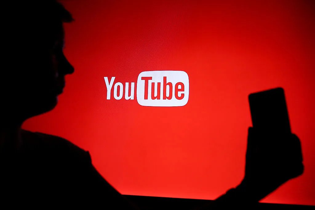 A silhouetted person in profile holds a phone in front of a red backdrop displaying the YouTube logo.