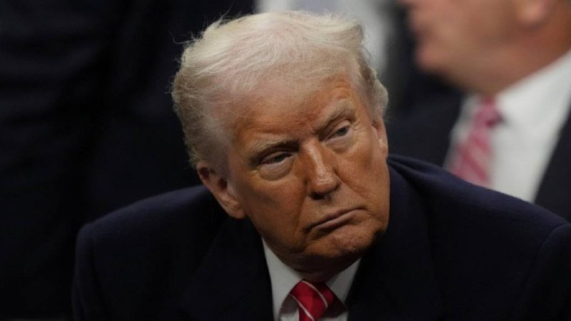 A man with blonde hair wearing a dark suit, white shirt, and red striped tie looks downward with a serious expression, with another suited figure blurred in the background.