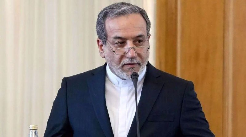 A gray-bearded man in a dark suit and white shirt speaks into a microphone against a wood-paneled background.