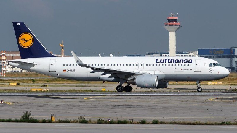 A white Lufthansa passenger jet taxis on a runway with an air traffic control tower and a Condor-branded building visible in the background.
