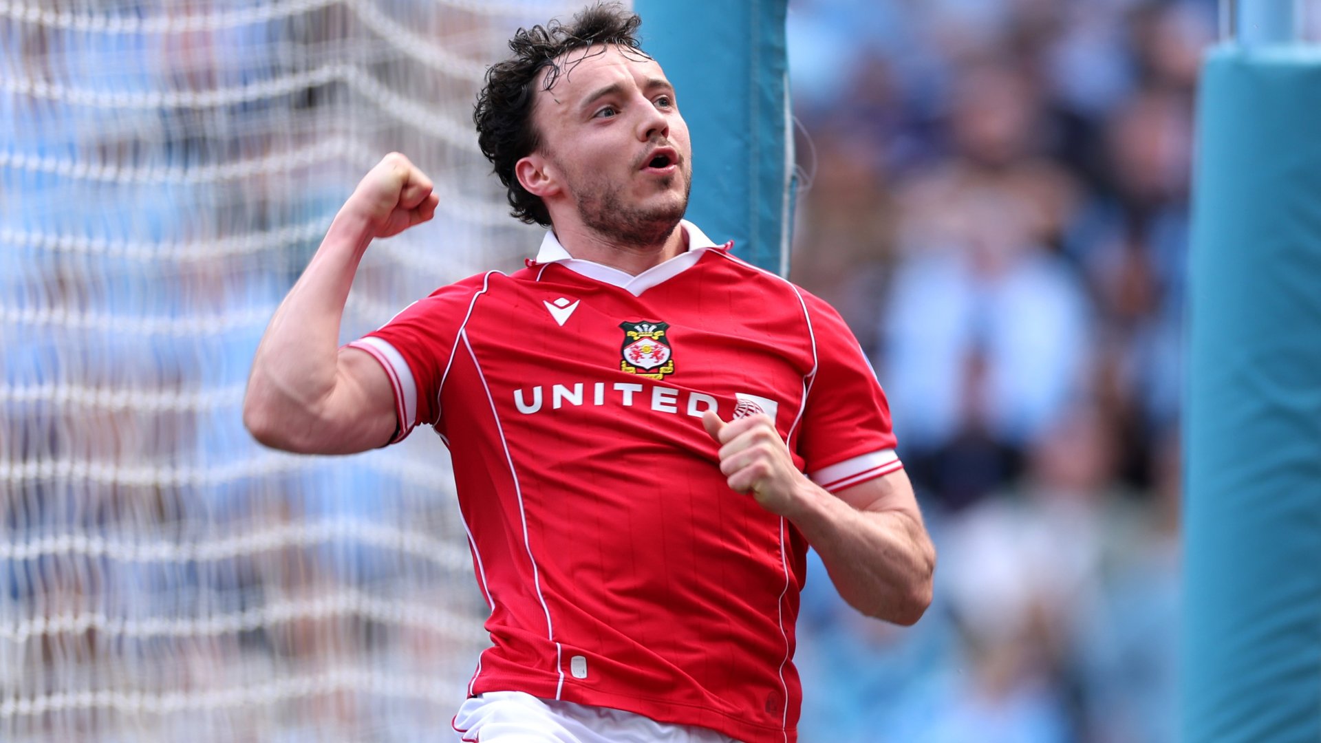 A male footballer in a red "UNITED" jersey celebrates with a raised fist in front of a goal net, with blurred spectators visible in the background.