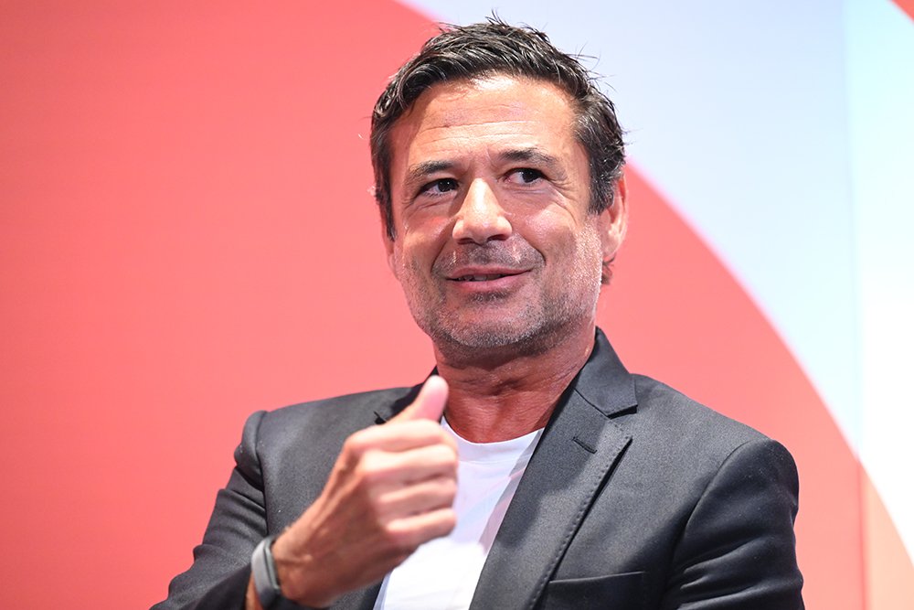 A man with dark hair in a black blazer and white t-shirt smiles while giving a thumbs-up against a red and white background.