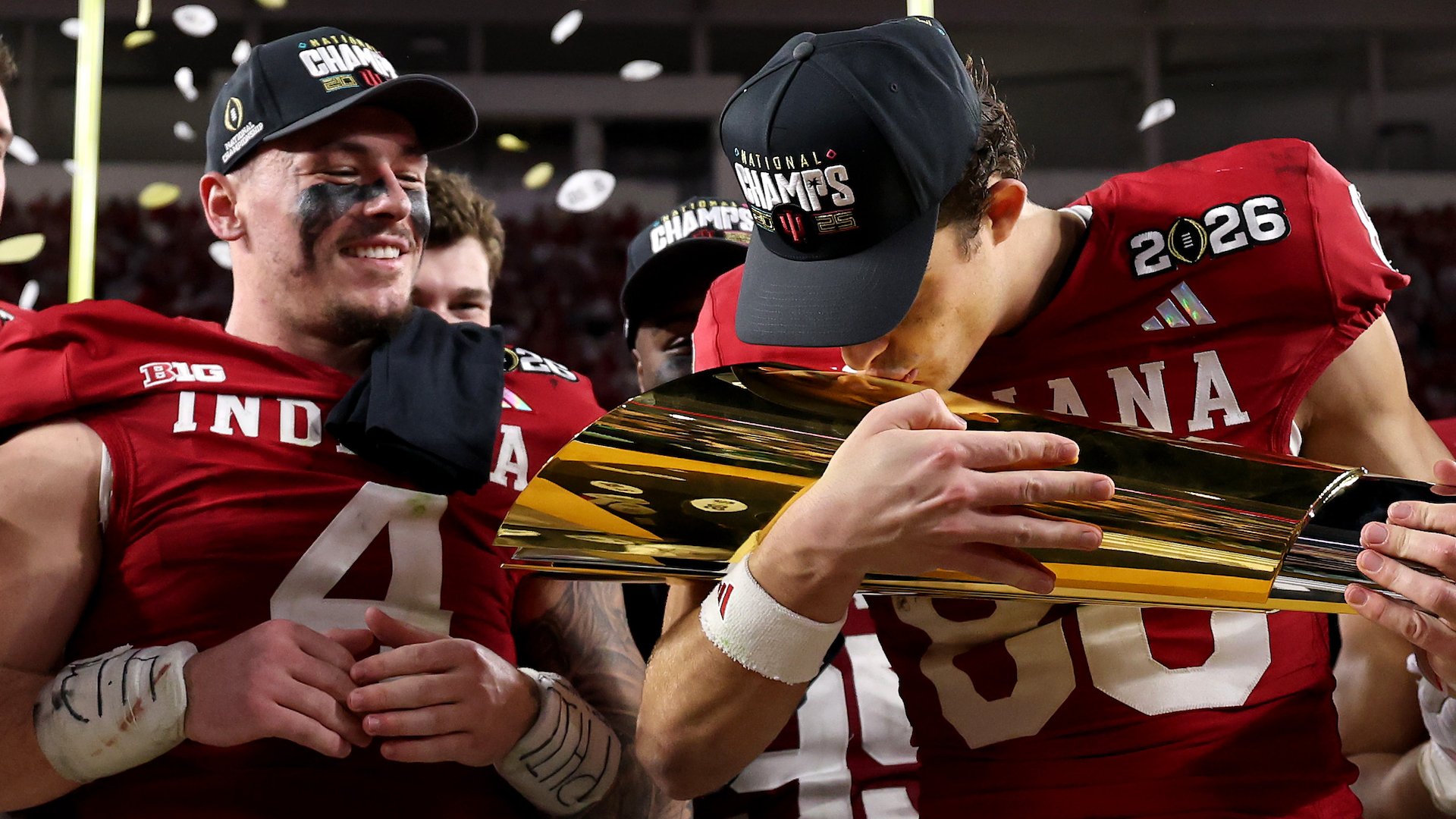 Football players in red "Indiana" jerseys and "National Champs" hats celebrate with a gold trophy as confetti falls in the background.