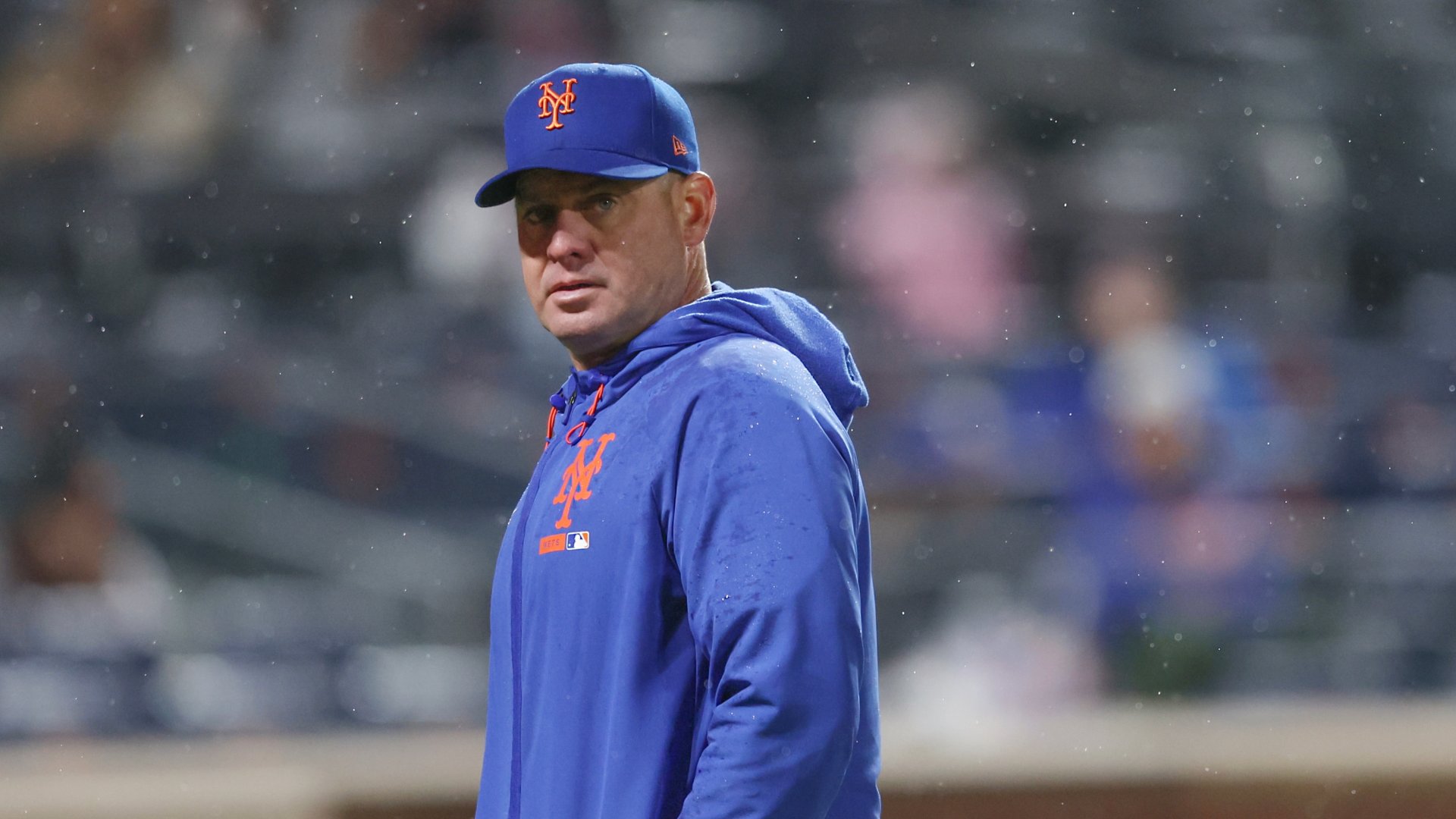 A man in a blue New York Mets cap and hoodie stands on a baseball field with rain falling, blurred spectators visible in the background.