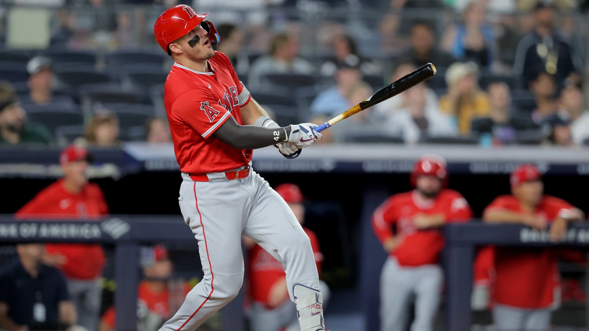 A baseball player in a red Angels jersey and helmet watches the ball after swinging his bat during a game, with teammates in the dugout and spectators visible in the background.