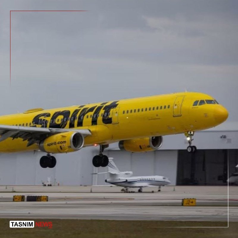 A yellow Spirit Airlines passenger jet with deployed landing gear flies low over an airport runway, with a smaller white private jet and hangar visible in the background.