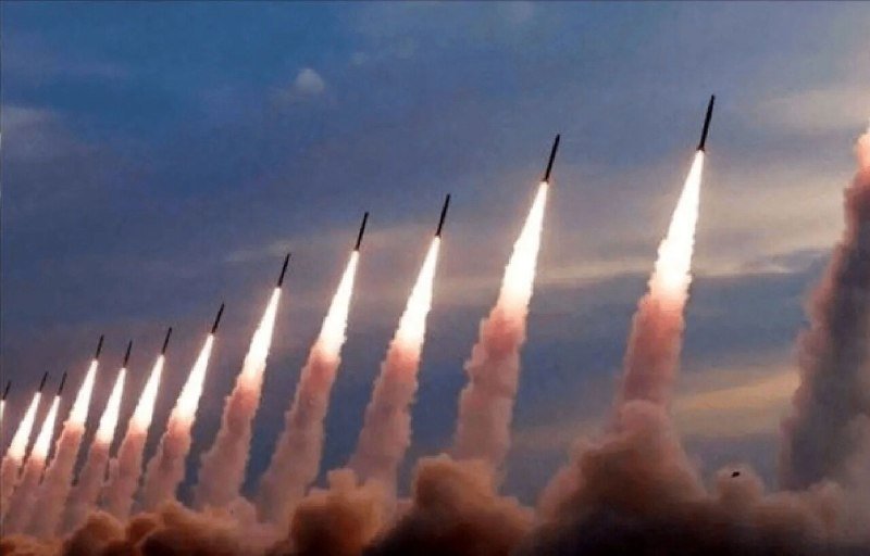 A row of missiles launches simultaneously into a cloudy sky, leaving bright white exhaust trails and billowing smoke below.
