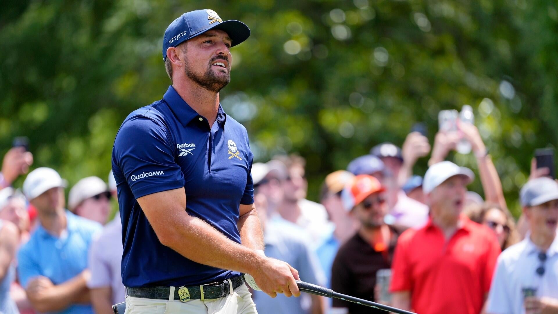 A bearded golfer in a navy Reebok polo and NetJets cap watches a shot's flight, holding a club while a crowd looks on.