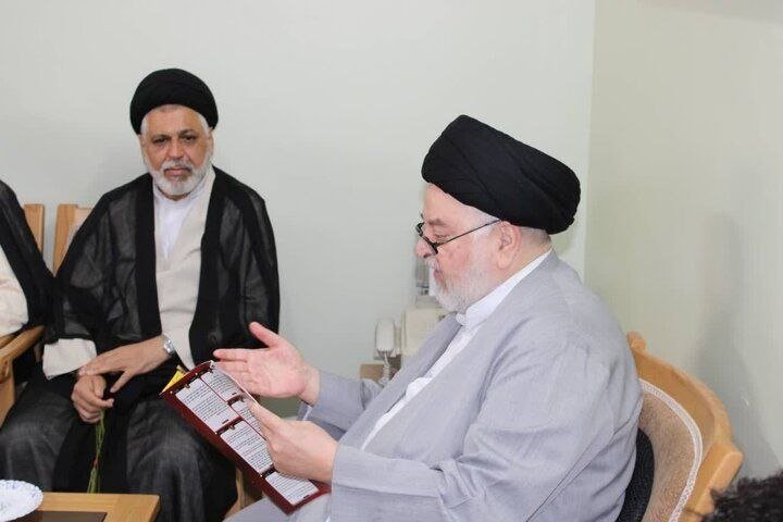 Two men wearing black turbans and traditional clerical attire sit indoors, one holding and gesturing toward an open booklet.