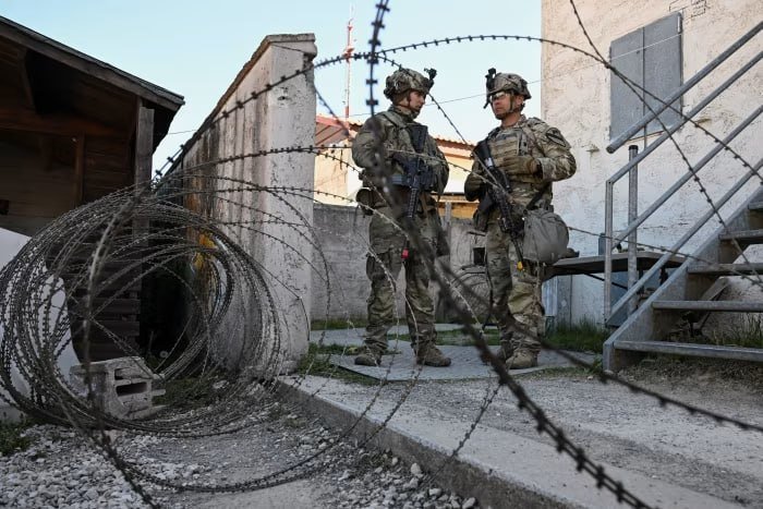 Two soldiers in camouflage uniforms and helmets stand behind coiled razor wire, holding rifles, near a building with metal stairs.