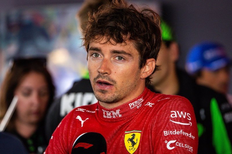 A man in a red Ferrari racing suit with sponsor logos, including AWS and Celsius, speaks into a microphone during an interview.