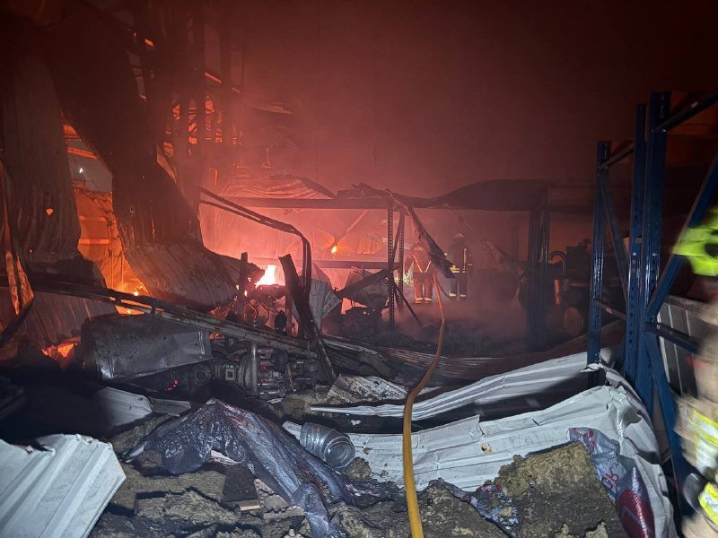 Firefighters in reflective gear stand amid the smoldering, collapsed wreckage of a structure illuminated by active flames and heavy smoke.