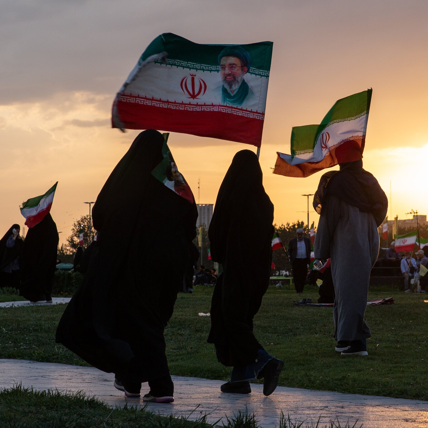 Women in black chadors and a man in gray robes walk along a paved path at sunset, carrying large Iranian flags, one featuring a portrait of a bearded man in a turban.
