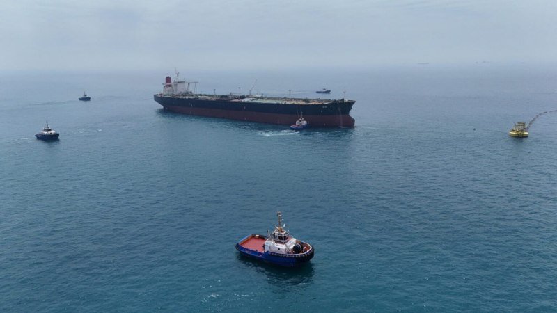Aerial view of a large cargo ship anchored offshore, accompanied by several smaller tugboats on calm, hazy waters.