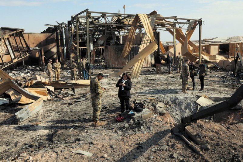 Soldiers in camouflage and individuals in civilian clothing inspect the wreckage of a destroyed building surrounded by scattered debris and twisted metal.