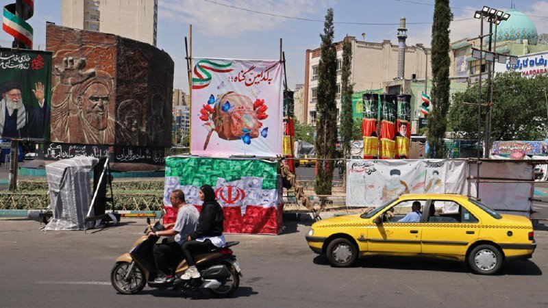 Two men ride a motorcycle and a driver navigates a yellow taxi past large political posters, banners, and a blue-domed mosque on a city street.
