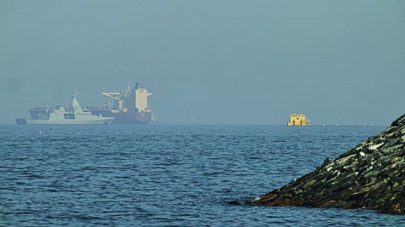 A gray naval warship and a large cargo vessel float in hazy open water on the left, with a small yellow boat to the right and a rocky shoreline in the foreground.