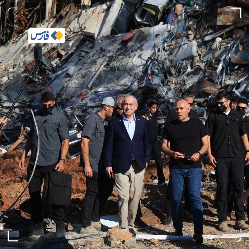 A group of men, including one in a blue blazer, walk past a large pile of twisted metal and concrete rubble.
