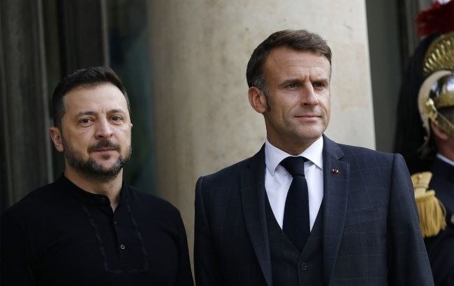 Two men stand side by side in dark suits, one in a black shirt and the other wearing a tie with a lapel pin, near a uniformed guard.