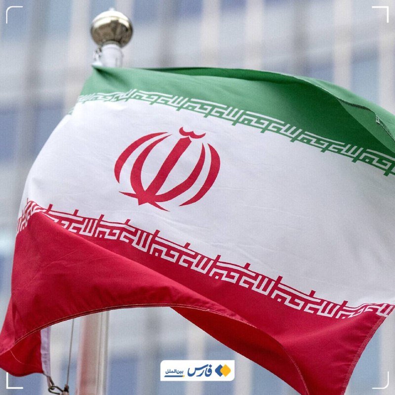 The flag of Iran, featuring green, white, and red horizontal stripes with a central red emblem, waves on a flagpole against a blurred background.