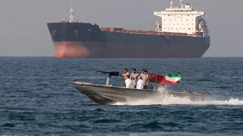 A small speedboat carrying several men and flying the Iranian flag speeds across the sea, with a large cargo ship visible in the background.