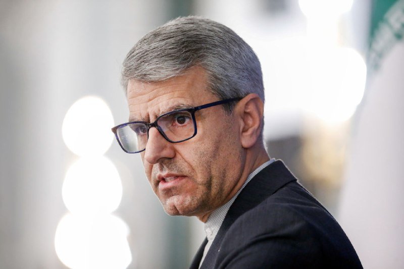 A gray-haired man wearing dark-framed glasses and a dark suit speaks during an indoor event.