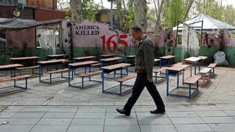 A man in a green jacket walks past rows of empty school desks arranged outdoors in front of a damaged wall reading "AMERICA KILLED 165."