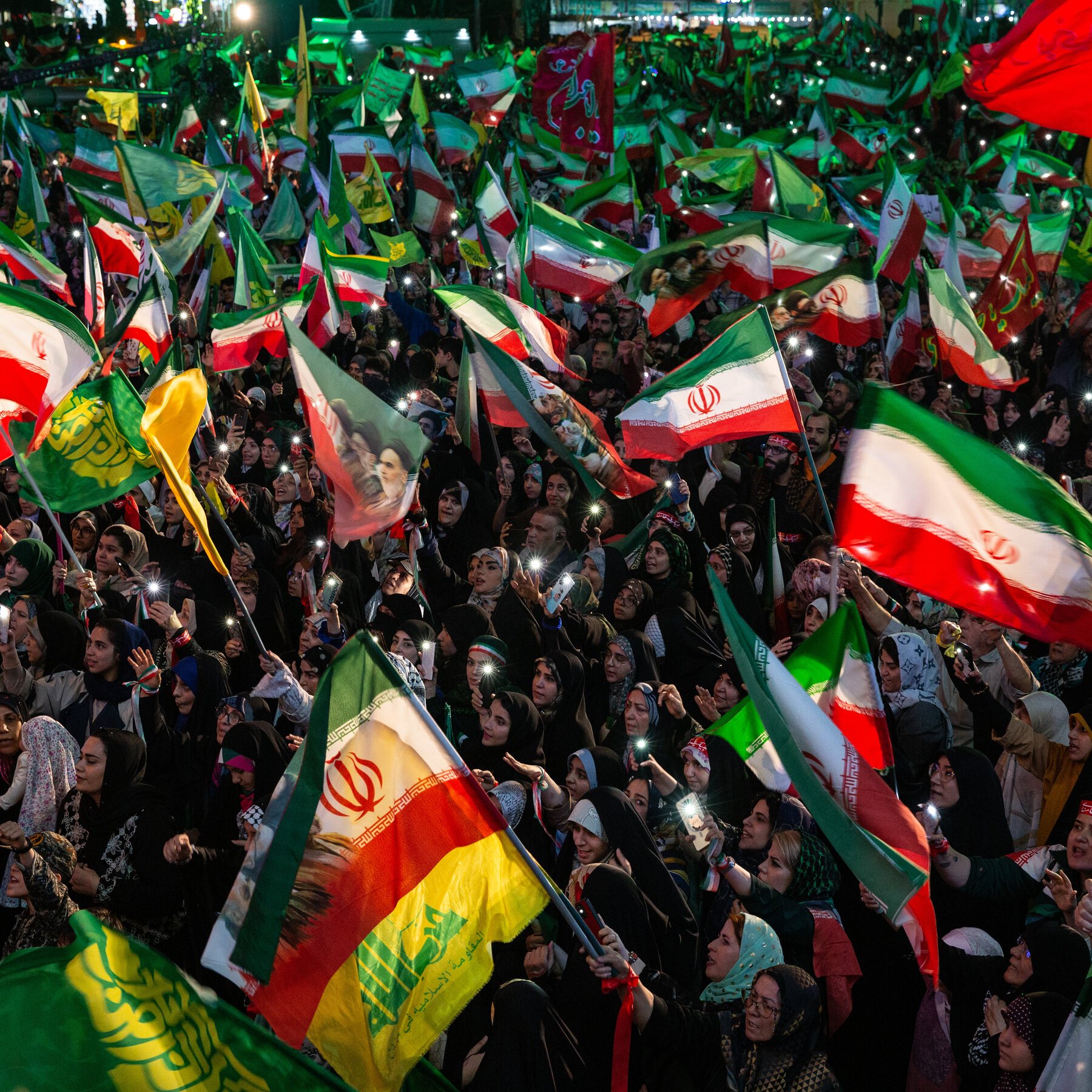 A nighttime crowd waves Iranian flags, green banners, and portrait flags while holding up lit phones.