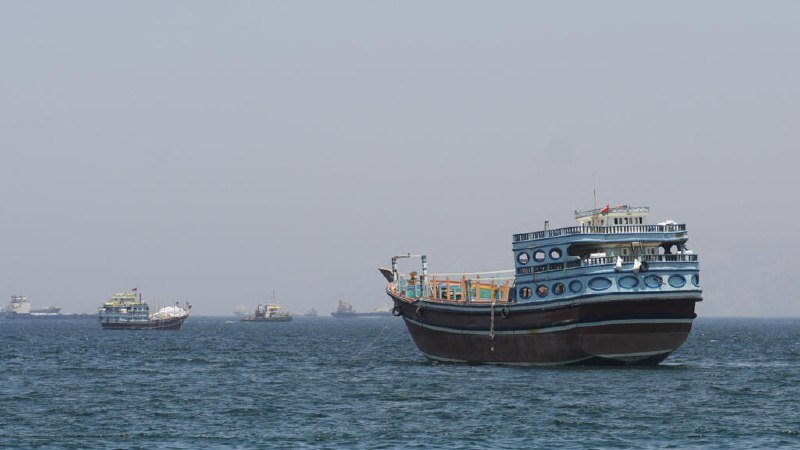 A large multi-deck blue and white boat floats in the foreground on open water, with several smaller vessels visible in the hazy distance under a pale sky.